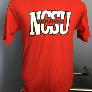 May include: Red t-shirt with a white and black graphic of the letters "NCSU" with the word "WOLFPACK" in black letters inside the "C".