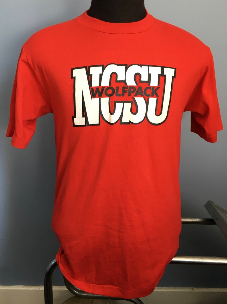 May include: Red t-shirt with a white and black graphic of the letters "NCSU" with the word "WOLFPACK" in black letters inside the "C".