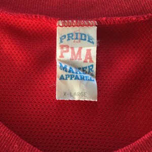 May include: A red, white, and blue tag with the text "Pride PMA Maker Apparel" and "X-Large" on a red shirt.