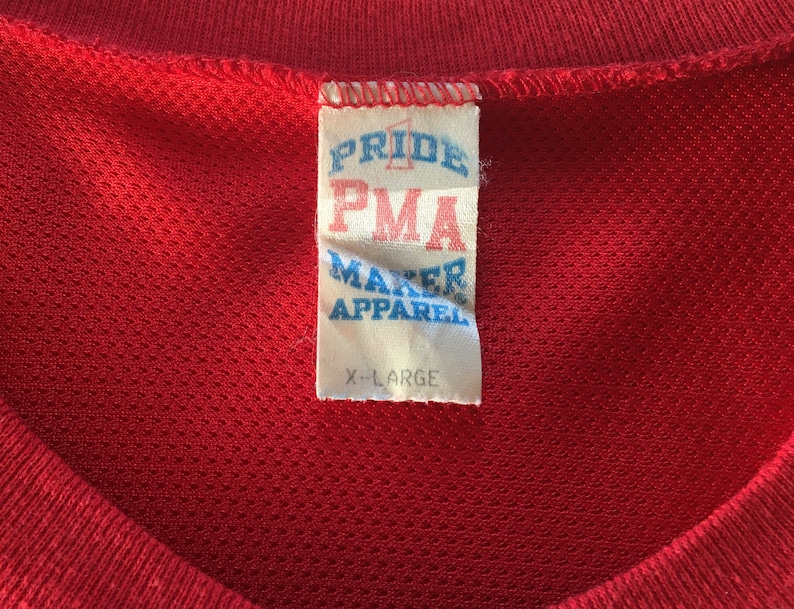 May include: A red, white, and blue tag with the text "Pride PMA Maker Apparel" and "X-Large" on a red shirt.