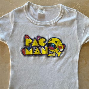 Può includere: T-shirt bianco con il logo di Pac-Man in giallo, blu, rosso e nero. La maglietta ha maniche corte e girocollo. La grafica di Pac-Man è centrata sulla parte anteriore della maglietta.