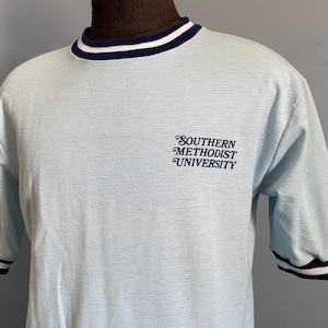 May include: A light blue short-sleeved t-shirt with a ribbed crew neckline and sleeve cuffs featuring dark blue and white stripes. The chest displays the text SOUTHERN METHODIST UNIVERSITY in a dark blue, stylised, cursive-inspired font.