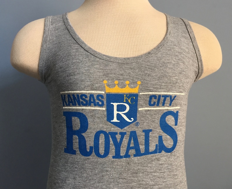 May include: Gray tank top with a blue and white Kansas City Royals logo. The logo features a crown, a shield with the letter "R" and the words "Kansas City Royals".