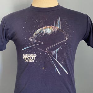 Könnte beinhalten: Marineblaues T-Shirt mit einer Grafik einer geodätischen Kugel, einem Sternenhintergrund und dem Text "Walt Disney World Epcot Center". Das Design umfasst weiße und blaue Linien und eine farbenfrohe Kugel.