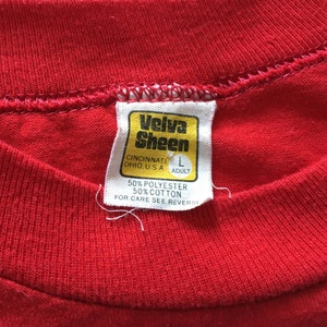 May include: A red Velva Sheen t-shirt tag with white stitching. The tag is yellow with black text that reads "Velva Sheen", "Cincinnati Ohio, U.S.A", "L Adult", "50% Polyester 50% Cotton", and "For Care See Reverse".