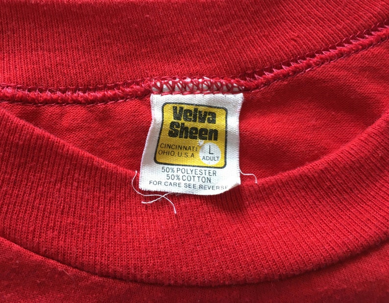 May include: A red Velva Sheen t-shirt tag with white stitching. The tag is yellow with black text that reads "Velva Sheen", "Cincinnati Ohio, U.S.A", "L Adult", "50% Polyester 50% Cotton", and "For Care See Reverse".
