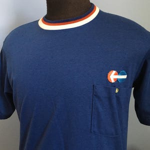 May include: A navy blue short-sleeved t-shirt with a pocket and a colorful embroidered logo. The shirt has a white, orange, and navy blue striped collar. A gold button is on the pocket.