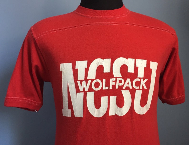 May include: Red t-shirt with a white graphic of the letters "NCSU" with the word "Wolfpack" in between the letters.