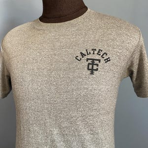 May include: A heather gray short-sleeve t-shirt featuring a crew neckline. The chest area displays the black text CALTECH arched above a black monogram logo consisting of the letters T and C intertwined in a stacked, block-style design.