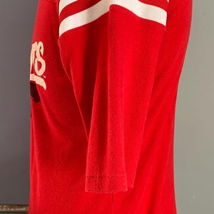May include: Red long-sleeve shirt with white stripes on the sleeves. The shirt has a logo on the front. The shirt is displayed on a mannequin.