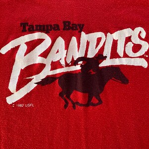 May include: Red fabric with the words "Tampa Bay Bandits" in white, with a black silhouette of a rider on a horse. The text "1982 USFL" is below the logo.