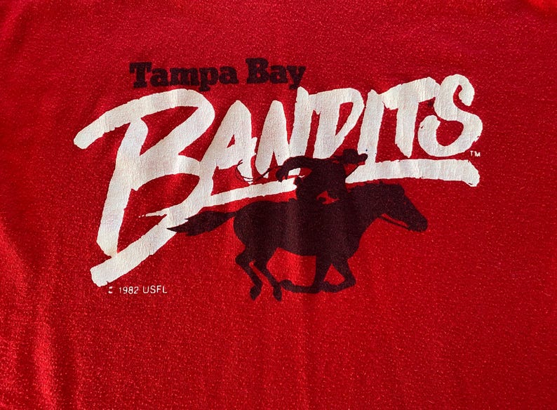 May include: Red fabric with the words "Tampa Bay Bandits" in white, with a black silhouette of a rider on a horse. The text "1982 USFL" is below the logo.