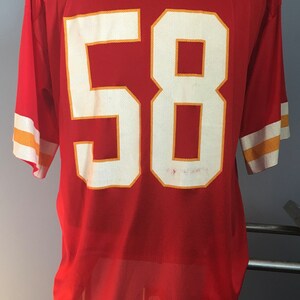 May include: Red football jersey with white number 58 outlined in gold. The jersey has white and gold stripes on the sleeves.