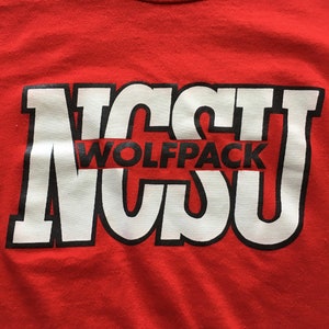 May include: Red t-shirt with a white and black graphic of the letters "NCSU" with the word "Wolfpack" in between the letters.