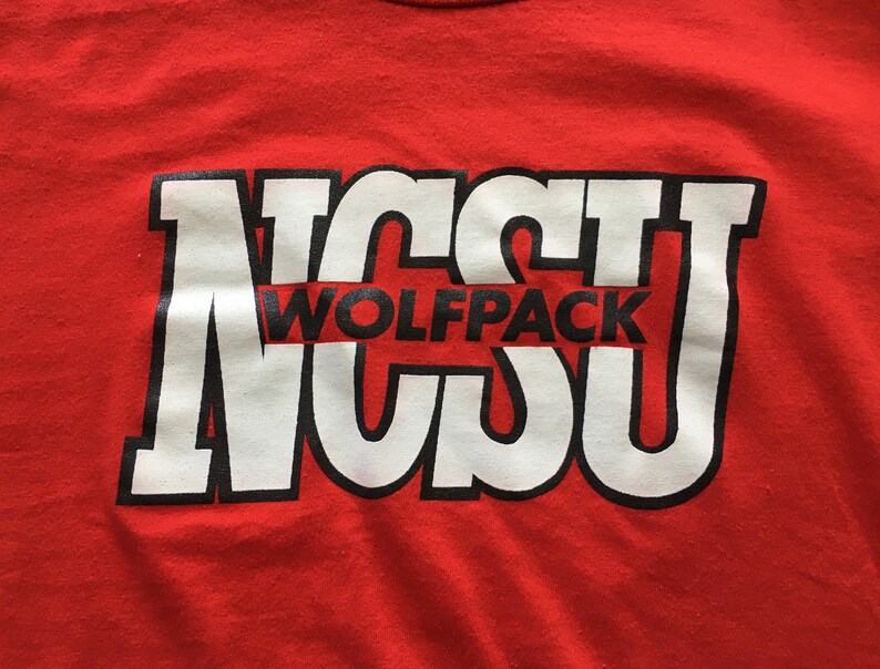 May include: Red t-shirt with a white and black graphic of the letters "NCSU" with the word "Wolfpack" in between the letters.