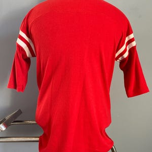 May include: A red three-quarter sleeve shirt with white stripes on the sleeves. The shirt is made of a soft material and has a classic athletic design. The shirt is displayed on a mannequin.