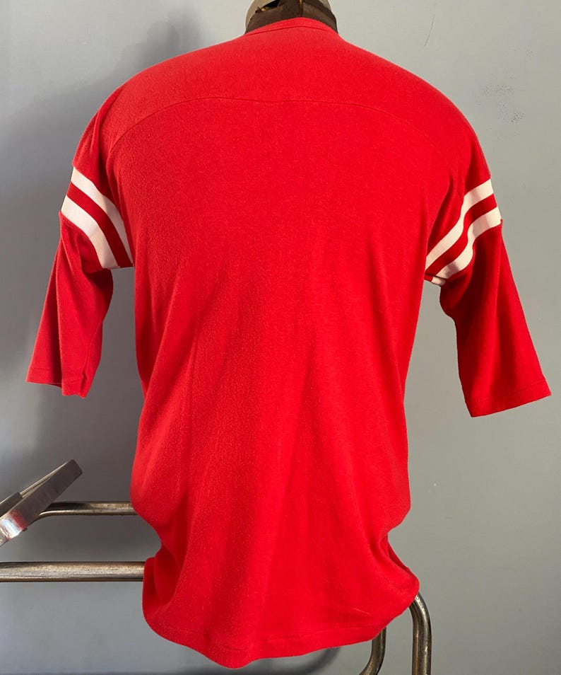 May include: A red three-quarter sleeve shirt with white stripes on the sleeves. The shirt is made of a soft material and has a classic athletic design. The shirt is displayed on a mannequin.