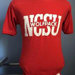 May include: Red t-shirt with white lettering that reads "NC State Wolfpack" in a retro style font. The shirt is a size M and is made in the USA.