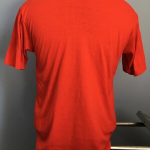 May include: A plain red short-sleeved t-shirt. The shirt is on a mannequin and is hanging on a metal rack.