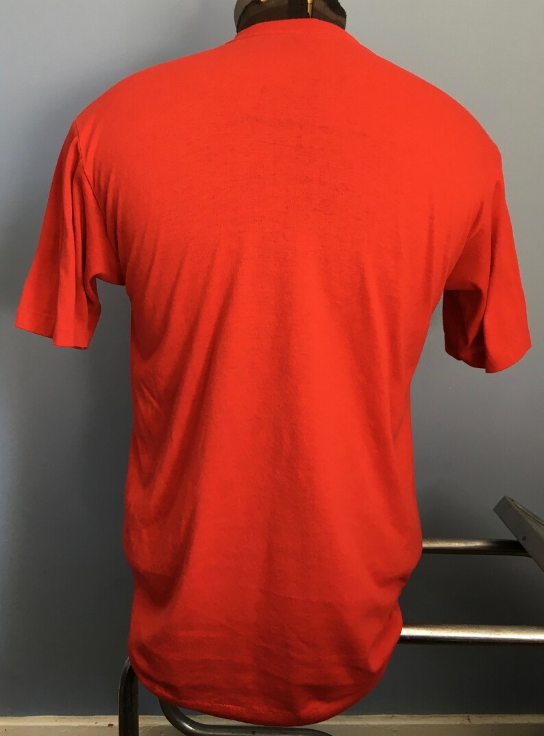 May include: A plain red short-sleeved t-shirt. The shirt is on a mannequin and is hanging on a metal rack.