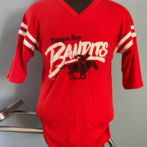 May include: Red vintage jersey-style shirt with white stripes on the sleeves. The shirt features the text "Tampa Bay Bandits" in white script with a black horse graphic. The shirt has a V-neck and three-quarter length sleeves.
