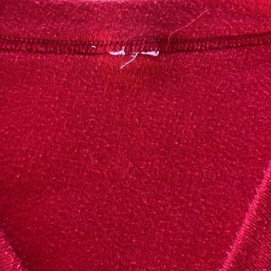 May include: Close-up of a red V-neck shirt. The fabric has a slightly textured appearance. The neckline is reinforced with a darker red stitching. A small white tag is visible near the neckline.