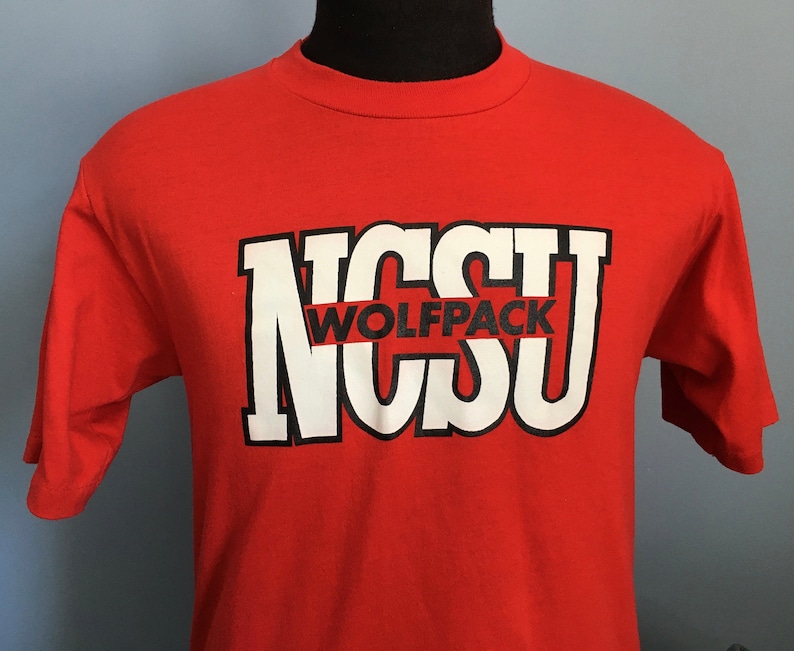 May include: Red t-shirt with a white graphic of the letters "NCSU" with the word "Wolfpack" in black letters across the middle of the letters.