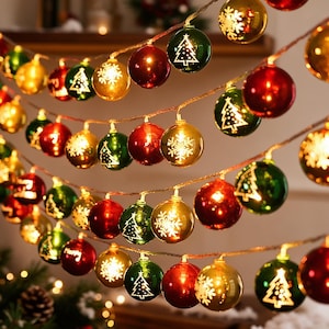 Pode incluir: Um cordão de luzes de Natal com ornamentos esféricos em vermelho, verde e dourado. As esferas são decoradas com desenhos de linhas brancas de árvores de Natal e flocos de neve, com algumas lâmpadas iluminadas por dentro.