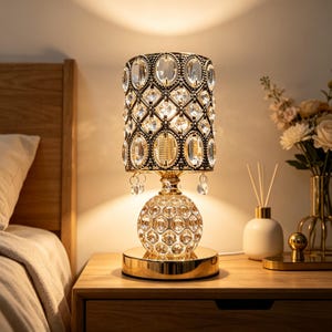 Może przedstawiać: Lampa stołowa z cylindrycznym kloszem o ciemnej metalowej ramie z wstawionymi przezroczystymi owalnymi kryształami. Podstawa to szklana kula o fasetowanej powierzchni, wsparta na polerowanym złotym metalowym stojaku i okrągłej podstawie.