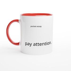 Può includere: Una tazza in ceramica bianca con interno rosso brillante e manico rosso abbinato. L'esterno presenta il testo nero pocket essay stampato vicino alla parte superiore e il testo nero pay attention. in caratteri più grandi verso il basso.