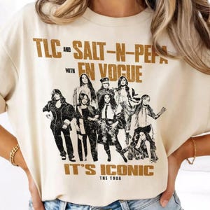 Può includere: Una t-shirt color crema con una stampa grafica marrone dall'aspetto vissuto dei gruppi musicali TLC, Salt-N-Pepa ed En Vogue. Il testo recita TLC AND SALT-N-PEPA WITH EN VOGUE IT'S ICONIC THE TOUR, raffigurando gli artisti in un ritratto di gruppo.