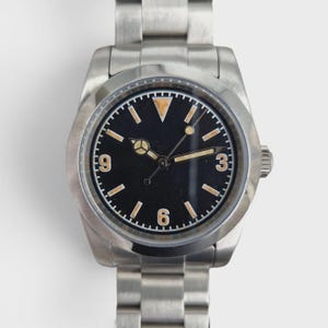May include: A stainless steel wristwatch with a black dial featuring a triangular marker at the twelve o'clock position, Arabic numerals at three, six, and nine, and baton-style hour markers. The watch includes a metal link bracelet and hands.