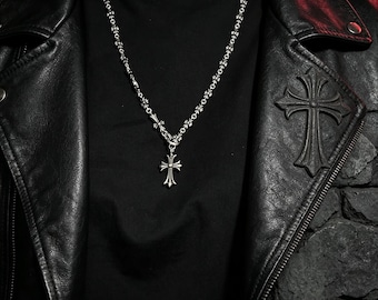 Puede incluir: Un collar de metal plateado con una cadena de eslabones ornamentados en forma de cruz y un colgante de cruz central. El collar se lleva sobre una camiseta negra lisa y una chaqueta de cuero negra que muestra un gran emblema de cruz en relieve.