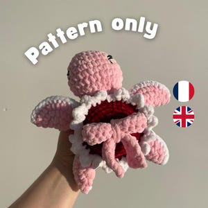 Peut inclure: Une peluche tortue au crochet avec une tête et des nageoires rose clair, une carapace rouge foncé bordée de blanc et un nœud rose clair au centre. Le texte "Pattern only" est visible au-dessus, accompagné d'icônes de drapeaux français et britannique.