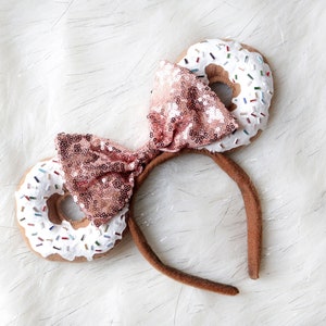 May include: A pair of donut-shaped headband ears with white frosting and colourful sprinkles. The headband is brown and has a pink sequined bow.