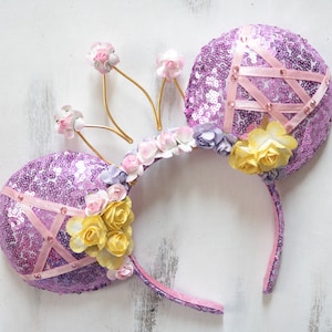 Pode incluir: Orelhas de Minnie Mouse de lantejoulas roxas com uma faixa de cabeça rosa e uma coroa dourada adornada com flores rosa, amarelas e brancas.
