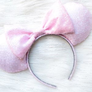 May include: Pink sparkly mouse ears headband with a large bow. The headband is made of a soft, plush material and is perfect for adding a touch of whimsy to any outfit.