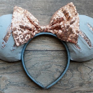 Pu&ograve; includere: Un paio di orecchie da topo in denim blu chiaro con un fiocco di paillettes oro rosa. Le orecchie hanno dettagli usurati e la fascia &egrave; nera.