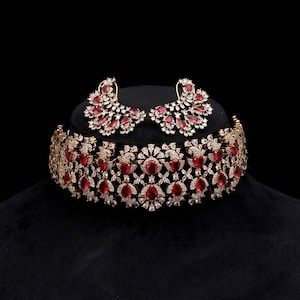May include: A wide choker necklace and a pair of matching earrings featuring a dense arrangement of clear white crystals and teardrop-shaped red stones. The jewelry set displays an intricate metalwork pattern with a symmetrical floral design.