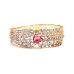 May include: A gold-toned hinged bangle bracelet featuring a central pear-shaped pink gemstone surrounded by numerous small, clear, sparkling stones arranged in a leaf-like pattern, with a lattice-work design on the inner band structure.
