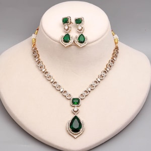 Può includere: Un set composto da collana e orecchini color oro con pietre verde scuro a forma di pera e rettangolari, circondate da piccoli cristalli trasparenti incastonati a pavé. La catena della collana presenta maglie a forma di infinito con cristalli.