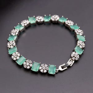 May include: A silver-toned bracelet featuring alternating rectangular mint green stones and circular clusters of small clear white stones. The bracelet is secured with a metal box clasp closure, creating a continuous circular link design.