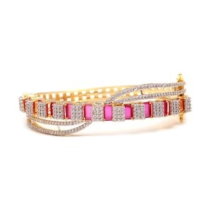 May include: A gold-toned hinged bangle bracelet featuring a row of rectangular pink stones alternating with square clusters of small clear white stones. The design includes curved, pavé-set clear stone accents on the top and bottom edges.
