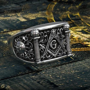 Op de afbeelding: Een zilverkleurige metalen ring met gedetailleerde reliëfsculpturen van vrijmetselaarssymbolen, waaronder een winkelhaak en passer met de letter G, twee zuilen met bollen en een zon-embleem op een donkere, verdiepte achtergrond.