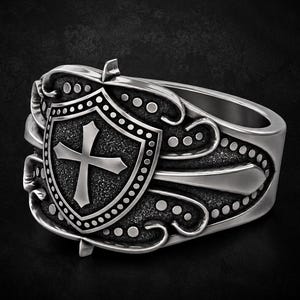 Könnte beinhalten: Ein silberfarbener Ring mit einem zentralen Schildelement, das mit einem Kreuz geprägt ist. Der Schild ist von kunstvollen Schnörkeln und einem gepunkteten Randmuster umgeben. Die Metalloberfläche ist poliert und weist abgedunkelte vertiefte Bereiche auf, um die detaillierte Reliefarbeit am Ringband hervorzuheben.