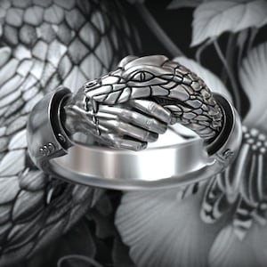 Könnte beinhalten: Ein silberfarbener Ring mit einem detaillierten skulpturalen Design einer menschlichen Hand, die den Kopf einer Schlange greift. Die Metalloberfläche hat ein poliertes Finish mit dunklen Vertiefungen, die die Schuppen und Finger betonen.