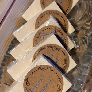 Peut inclure: Une pile de barres rectangulaires de savon blanc cassé enveloppées dans du plastique transparent. Chaque barre comporte une étiquette ronde en papier brun avec une bordure florale et le texte bleu manuscrit Coconut Patchouli.