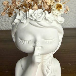 May include: A white ceramic planter shaped like a head with closed eyes and a finger held to the lips in a shushing gesture. The figure wears a crown of sculpted flowers and has a braided hairstyle. Dried beige flowers are arranged inside the top.