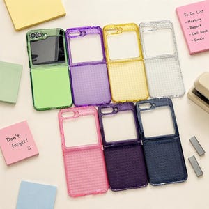 May include: A collection of seven foldable phone cases in various colours including green, purple, yellow, clear, pink, dark purple, and dark blue. Each case features a grid-textured lower section and a clear upper frame with camera cutouts.