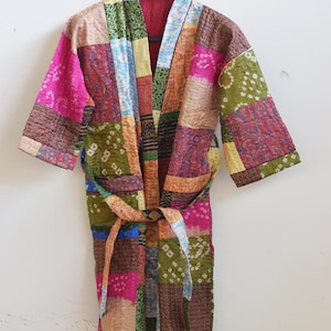 Pode incluir: Um roupão até o joelho com um design de patchwork composto por vários quadrados de tecido em cores vibrantes, incluindo rosa, verde, marrom e azul. A peça possui mangas compridas, gola larga e um cinto de tecido a condizer.